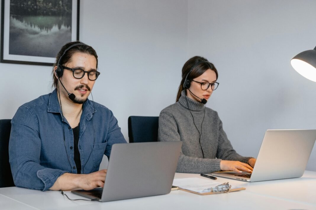 Two customer service representatives wearing headsets and working on laptops in a modern office setting. The man on the left is wearing a denim shirt and glasses, while the woman on the right is wearing a gray turtleneck sweater and glasses.