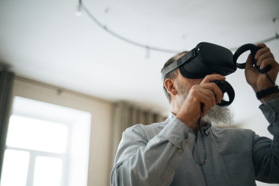 An elderly man with a gray beard wearing a virtual reality (VR) headset and holding VR controllers, engaging in a virtual experience in a well lit indoor setting.